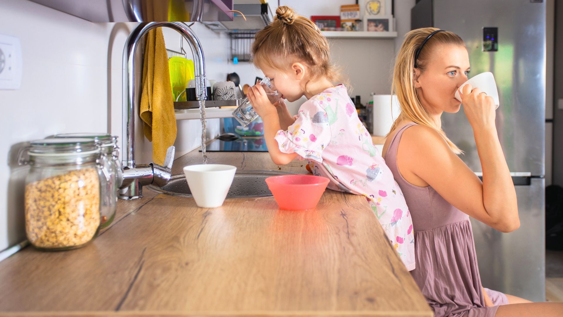 Mother and young child drinking water from cups in a kitchen, highlighting concerns about tap water safety for babies