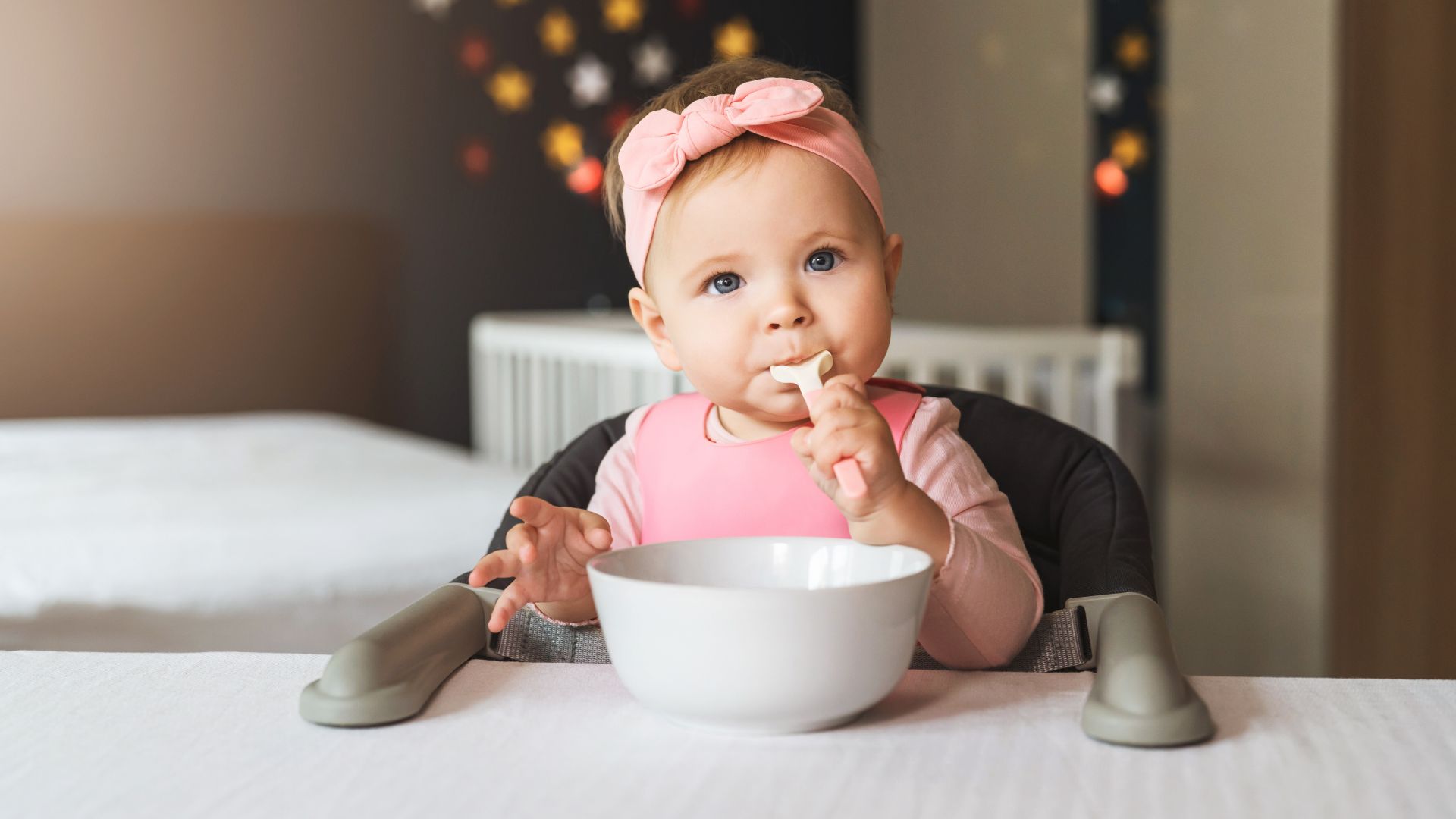 Spoon Grasp Development for Toddlers Baby learning to eat independently while sitting in a high chair, holding a spoon and reaching toward a bowl on the table.