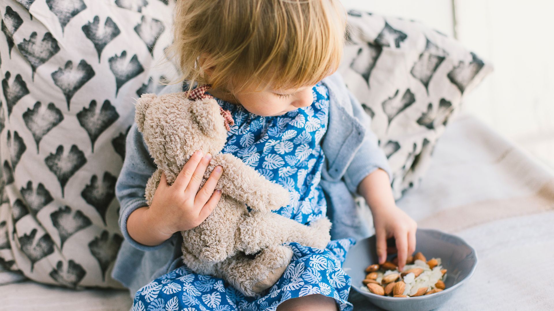 Toddler sitting on a couch holding a stuffed toy while reaching for nuts from a bowl