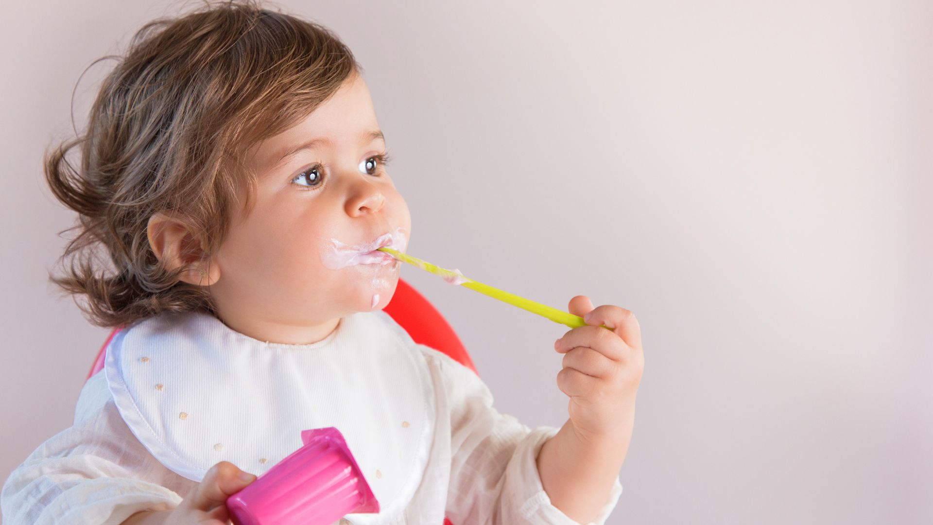 Baby sitting in a high chair eating yogurt with a spoon, relating to when babies can safely have yogurt